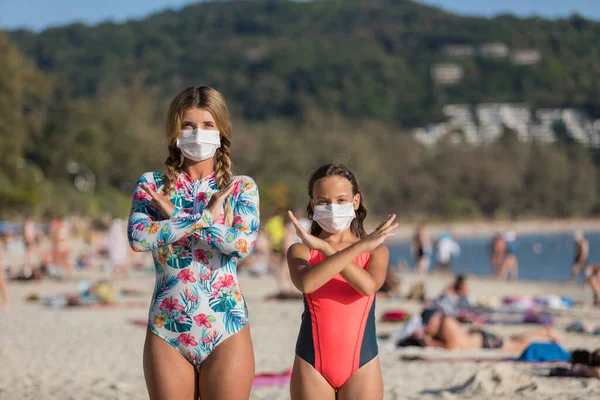 Yüzlerinde koruyucu cerrahi maske takmış bir kadın ve bir çocuk sahilde mayo giymiş duruyor. Çin koronavirüs hastalığı COVID-19 tehlikelidir..