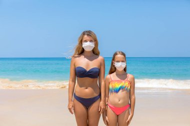 Yüzlerinde koruyucu cerrahi maske takmış bir kadın ve bir çocuk sahilde mayo giymiş duruyor. Çin koronavirüs hastalığı COVID-19 tehlikelidir..