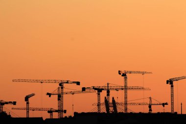 Gün batımında yüksek kule vinçlerinin siluetleri şehir binalarının üzerinde