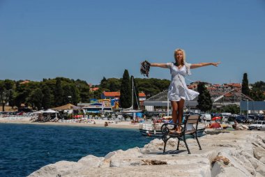 Elle İskele'nın banka deniz kenarında geniş açık mavi elbiseli bir genç sarışın bir kadın duruyor.