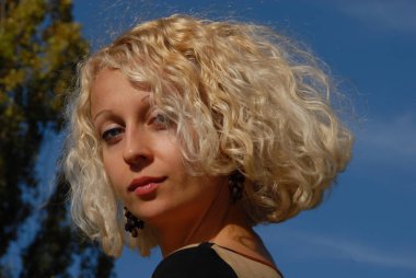 Sarı kıvırcık saçlı ve mavi gözleri mavi gökyüzünün altında olan güzel bir kadın portresi.