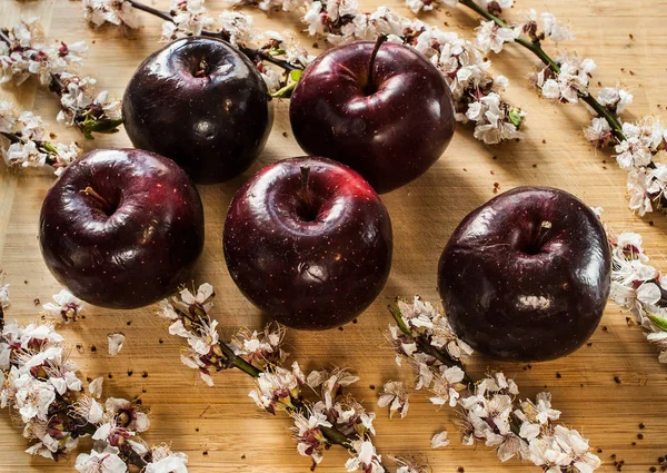 Five crimson ripe apples and apple tree branches with flowers lay on a wooden cutting board.