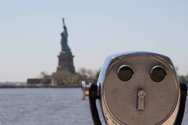 Tourist binoculars on Ellis Island with Statue of Liberty visible in the blurred background. There is a blue sky and water visible. The metal shows age and is burnished. clipart