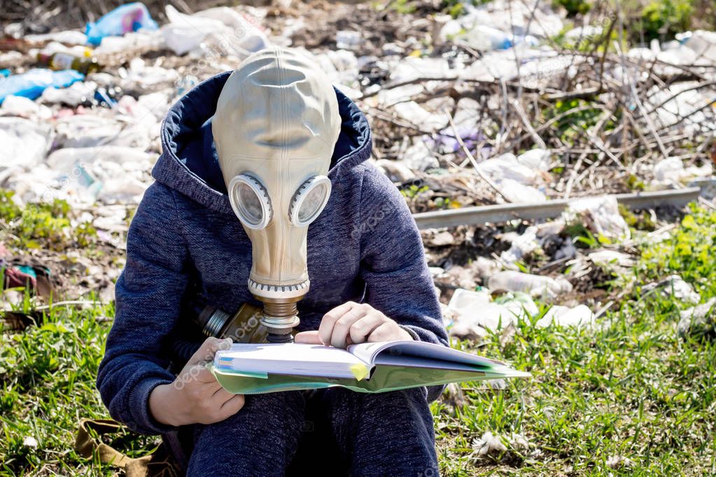Boy in  gas mask reads  book on the background of  garbage. Ecol