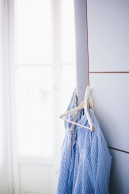 Erkeklerin gömlekleri dolaptan sarkıyor, odanın pencerelerinden içeri ışık giriyor.