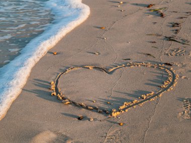 Deniz kıyısı, kuma çizilen kalp dalgalardan, gün batımından silinir.