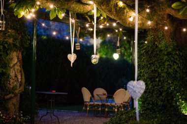 Geniş manolya ağacıyla dolu villa bahçesi. Dallarından sarkan bir sürü fener ve elektrik lambası var. Düğün partisi ya da diğer kutlamalar.