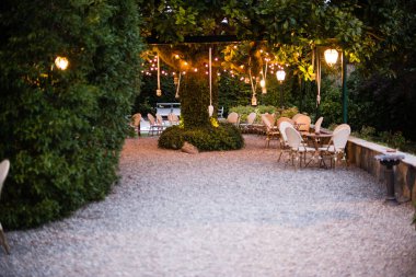 Geniş manolya ağacıyla dolu villa bahçesi. Dallarından sarkan bir sürü fener ve elektrik lambası var. Düğün partisi ya da diğer kutlamalar.