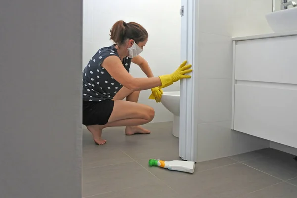 Adult woman with face mask and working rubber gloves cleaning and sanitizing home rest room using disinfectant bleach cleaner to kill and prevent coronavirus spread. Real people. Copy space.