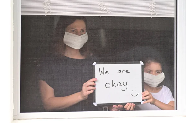 Mother and daughter (age 10)  in self isolation wearing a medical face mask looking outside home window holding a sign reading: We are okay to relatives.