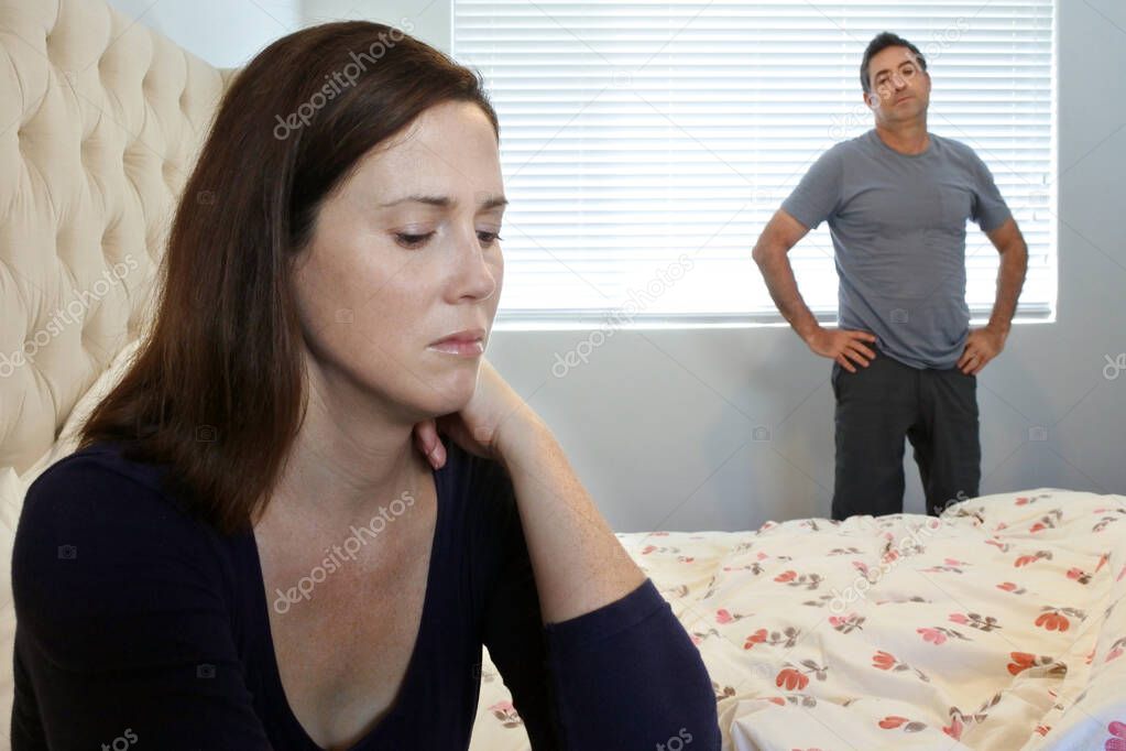 Upset couple in home bedroom. woman sitting on bed in forground and man standing in the  background.