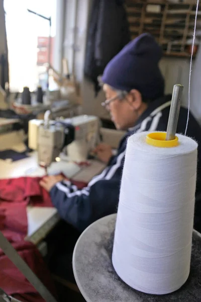 Professional tailor (age 70) using sewing machine to stitch fabrics in traditional tailors shop. Real people