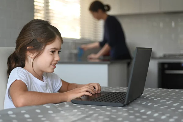 母親が台所を掃除している間 若い女の子 10歳 は自宅でノートパソコンを使用しています 本物の人間だ スペースのコピー — ストック写真
