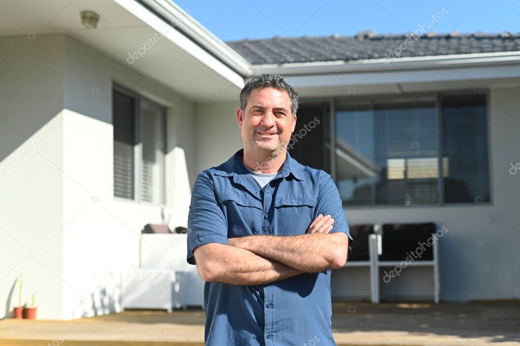 Happy adult man (age 35-45) standing in front of his new home. Buy, sell, real estate, property, home insurance, mortgage, bank loans and housing market concept. Real people. Copy space
