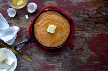Whole Yellow Cornbread  with butter in cast iron skillet and red pot holder on rustic wood table top view clipart