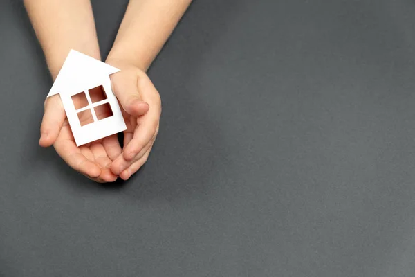 Vista superior en manos de niños sosteniendo la casa de papel blanco sobre fondo gris. Casa familiar y concepto de bienes raíces. Colocación plana, espacio de copia —  Fotos de Stock