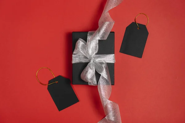 Gift box wrapped in black paper with silver ribbon and price tags on bright red background. Copy space and top view. Black friday box gift present isolated.