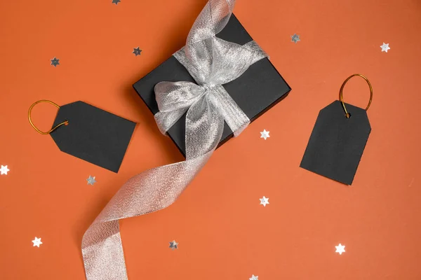 Black friday sale concept. Black paper boxes with silver ribbon, price tags and stars confetti on bright orange background. Flat lay, top view, copy space