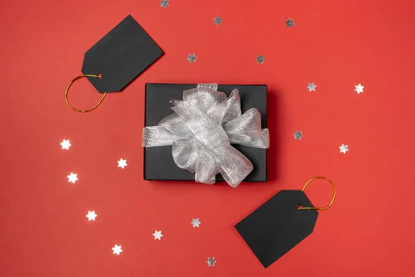 Coffret cadeau enveloppé dans du papier noir avec ruban argenté, étoiles confettis et étiquettes de prix sur fond rouge vif. Copier l'espace et la vue du dessus. Cadeau cadeau boîte vendredi noir isolé . — Photo