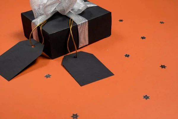 Dárková krabička zabalená v černém papíru se stříbrnou stuhou, hvězdami konfety a cenovkami na jasně oranžovém pozadí. Kopírovat prostor a pohled shora. Černá páteční box dárek prezentovat izolované. — Stock fotografie