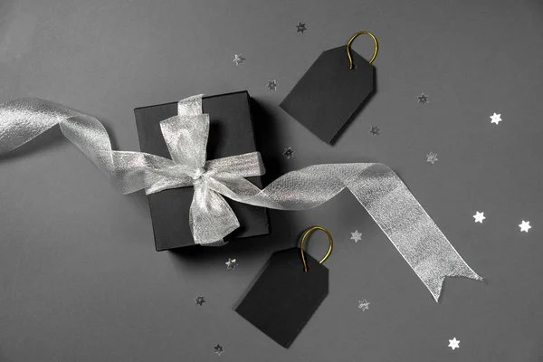 Gift box wrapped in black paper with silver ribbon, stars confetti and price tags on gray background. Copy space and top view. Black friday box gift present isolated.