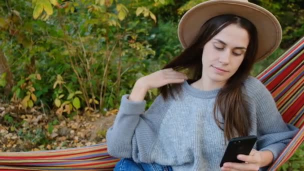 年轻女人挂在吊床上 被社交媒体或游戏应用程序分散注意力 穿着灰色毛衣和帽子 动作缓慢 社交媒体影响人们或内容制造者在自然中放松的观念 — 图库视频影像