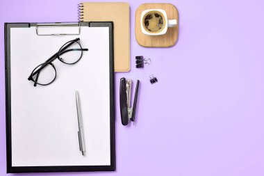 Üst kattaki ofis masasında yatıyordu. Çalışma alanı boş klip tahtası, not defteri, gözlükler, ofis malzemeleri, zımba, mor arka planda kalem ve kahve fincanı. Günlük kavramı