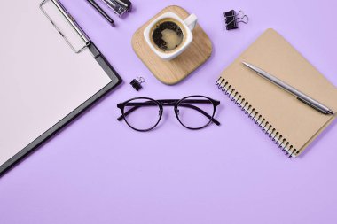 Çalışma alanını boş klip tahtası, ofis malzemeleri, kalem, kahve fincanı, not defteri, zımba ve mor arka planda gözlük ile düzenle. Düz konum, üst görünüm, kopyalama alanı