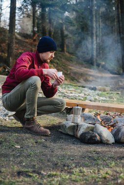 Genç kaşif erkeğin dağlarda sıcak bir içecek içerken, şenlik ateşinin yanında otururken, yürüyüş yaptıktan sonra dinlenirken çekilmiş açık hava çekimi. Mavi şapkalı gezgin, yürüyüşten sonra elinde bir kupa çay tutuyor..