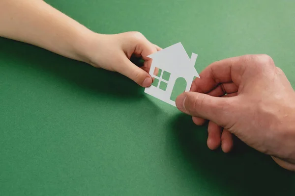 Primer plano de manos masculinas e infantiles sosteniendo la casa de papel blanco sobre fondo verde. Casa familiar y concepto de bienes raíces. Colocación plana, espacio de copia —  Fotos de Stock