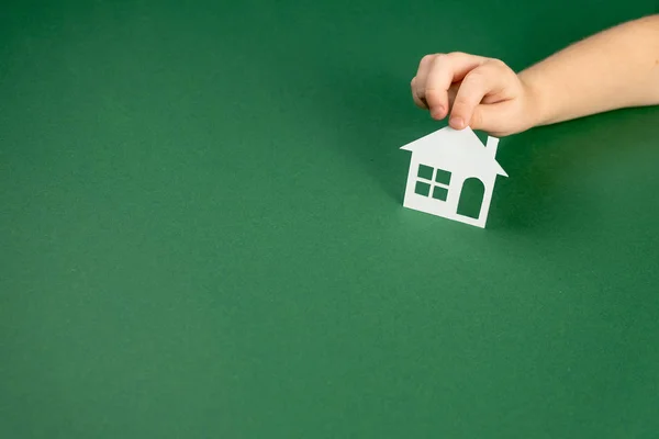 Widok z góry na ręce dziecka trzymając biały papierowy dom na zielonym tle. Dom rodzinny i koncepcja nieruchomości. Płaskie ułożenie, przestrzeń kopiowania — Zdjęcie stockowe