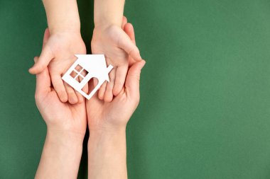 Close up of female and child hands holding white paper house on green background. Family home and real estate concept. Flat lay, copy space clipart