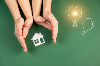 Family home and real estate concept. Close up of female and child hands holding white paper house and light bulb image on green background. Flat lay, copy space clipart