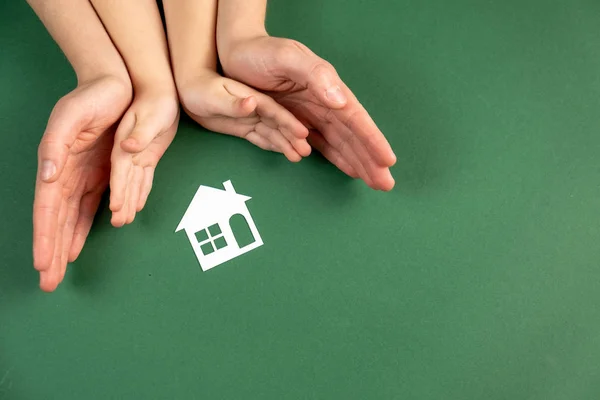 Casa de família e conceito imobiliário. Close up de mãos femininas e infantis segurando casa de papel branco e coração no fundo verde. Flat lay, espaço de cópia — Fotografia de Stock