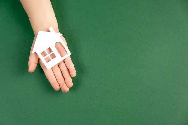 Visão superior na mão da criança segurando a casa de papel branco no fundo verde. Casa de família e conceito imobiliário. Flat lay, espaço de cópia — Fotografia de Stock