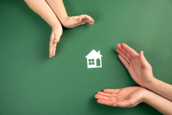 Családi ház és ingatlan koncepció. Közelkép a női és a gyermek kezét tartó fehér papírház és a szív zöld háttérrel. Lapos fektetés, fénymásolás — Stock Fotó