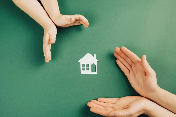 Primer plano de manos de mujeres y niños sosteniendo la casa de papel blanco y el corazón sobre fondo verde. Casa familiar y concepto de bienes raíces. Colocación plana, espacio de copia —  Fotos de Stock