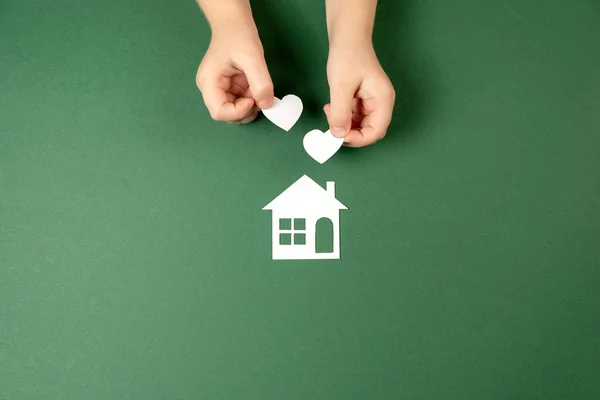 Mãos de criança segurando casa de papel branco e coração em fundo verde. Casa de família e conceito imobiliário. Flat lay, espaço de cópia — Fotografia de Stock