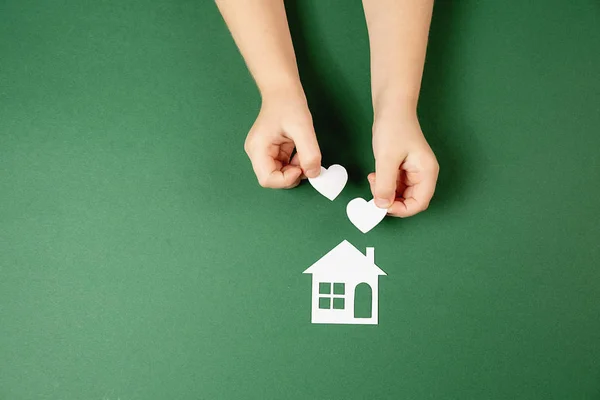 Casa de família e conceito imobiliário. Close up de mãos de criança segurando casa de papel branco e coração no fundo verde. Flat lay, espaço de cópia — Fotografia de Stock