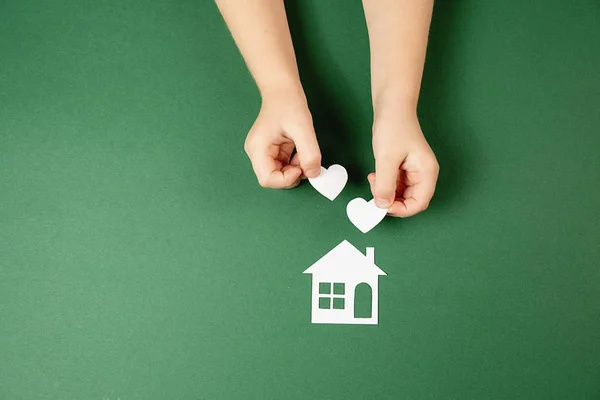 Casa di famiglia e concetto immobiliare. Primo piano di mani di bambino che tengono la casa di carta bianca e il cuore su sfondo verde. Posa piatta, spazio di copia — Foto Stock