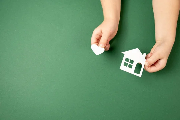 Mãos de criança segurando casa de papel branco e coração em fundo verde. Casa de família e conceito imobiliário. Flat lay, espaço de cópia — Fotografia de Stock