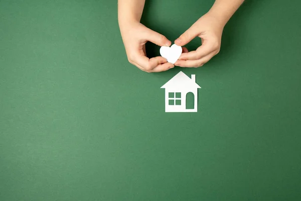 Casa de família e conceito imobiliário. Close up de mãos de criança segurando casa de papel branco e coração no fundo verde. Flat lay, espaço de cópia — Fotografia de Stock