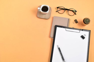 Üst kattaki ofis masasında yatıyordu. Çalışma alanı boş klip tahtası, ofis malzemeleri, kalem, kaktüs, ahşap bir stantta kahve fincanı ve turuncu arka planda gözlük. Günlük kavramı