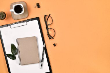 Ofis masası çalışma alanı boş klip tahtası, ofis malzemeleri, kalem, kaktüs, yeşil yaprak, ahşap bir stantta kahve fincanı ve turuncu arka planda gözlük. Düz konum, üst görünüm blog konsepti