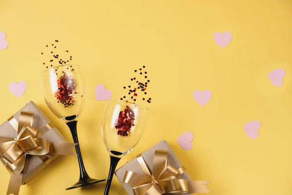 Fondo del Día de San Valentín. Dos copas de vino tintineo, varias cajas de regalo hechas a mano decoradas con confeti corazón rojo sobre fondo amarillo. Concepto de San Valentín. Plano, vista superior, espacio para copiar — Foto de Stock