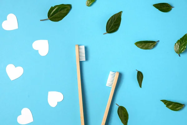 Dos cepillos de dientes ecológicos de bambú de madera, hoja verde, corazón blanco sobre fondo azul. Cuidado dental, ecológico, reutilización y concepto de día de San Valentín. Plano, vista superior, espacio para copiar — Foto de Stock