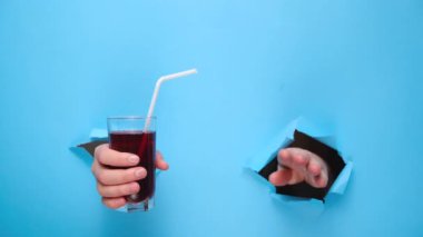 Kadın elinin, yırtık mavi kağıt duvardan geçen bir kamışla bir bardak taze kırmızı meyve suyu tutarken yakın plan. Meyve içecekleri ve sağlıklı beslenme. El işareti. Duvarda delik var. Jest gibi