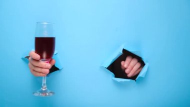 Kadının elinde bir kadeh kırmızı şarap tutuyor ve yırtık mavi kağıt bir duvar işareti gösteriyor. Sevgililer günü ve şarap barı, şaraphane, şarap tatma konsepti. El işareti. Duvarda delik var. Jest gibi