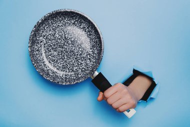 Unrecognizable female holding Frying pan through torn paper blue background. Kitchen utensils concept. Copy space for advertisement clipart