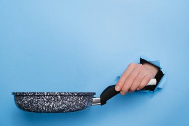 Yırtık kağıt mavisi arka planda dişi tavayı tutuyor. Mutfak aletleri konsepti. Reklam için boşluk kopyala
